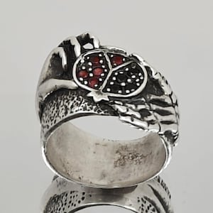 May include: A silver ring with a detailed design. The ring features a central design with red and black stones. The band has a textured pattern. The ring is displayed on a reflective surface.