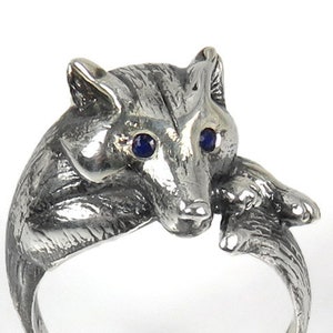 Könnte beinhalten: Ein silberner Ring mit einem detaillierten Fuchsdesign. Der Fuchs hat blaue Augen und ist mit seinen Pfoten unter dem Kinn zusammengerollt.