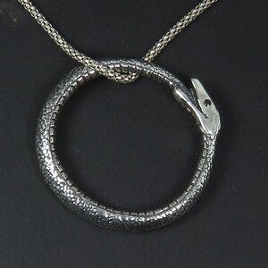 Op de afbeelding: Een zilveren ketting met een slangenhanger. De slang is opgerold tot een cirkel en zijn kop is naar voren gericht. De ketting is gemaakt van een delicate ketting.