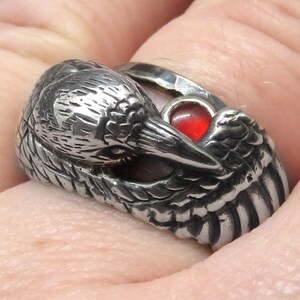 May include: A silver ring with a detailed bird design and a red gemstone in the eye. The bird's wings are spread out and the ring is worn on a finger.