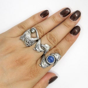 May include: A hand wearing three silver rings. The rings feature a dolphin design, a heart with a pearl, and a dolphin with a blue stone.