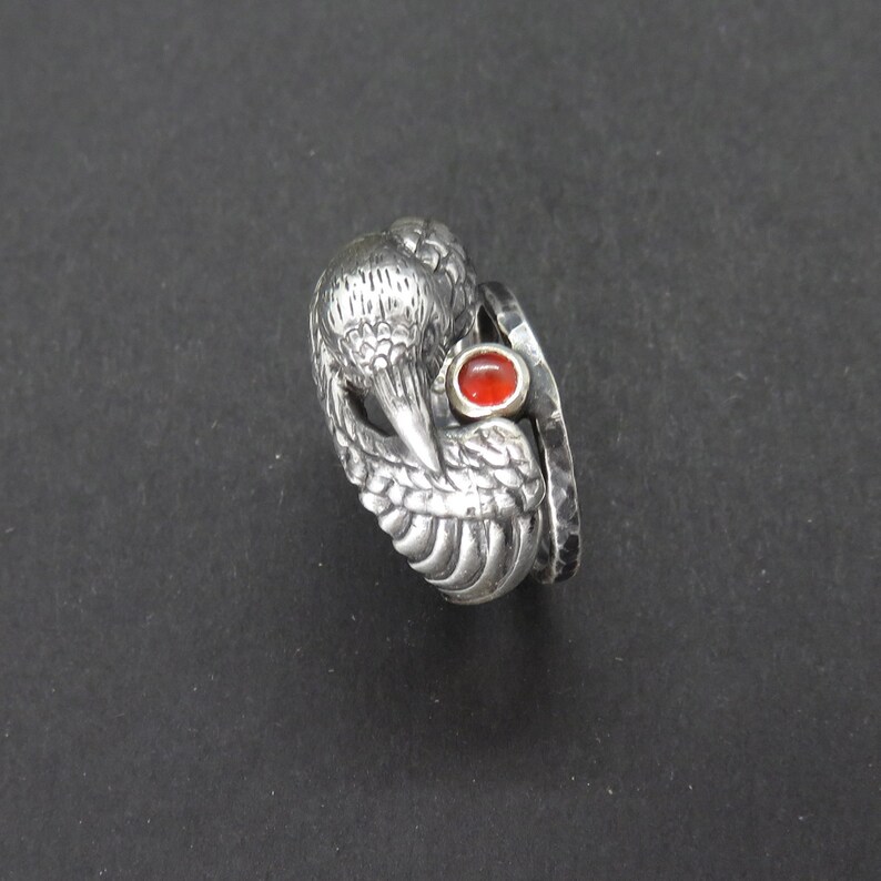 May include: A silver ring with a detailed bird design and a red gemstone. The ring is made of two bands, one with a textured finish and the other with a smooth finish.