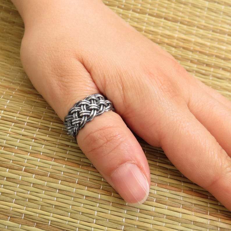 May include: A silver ring with a braided design. The ring is worn on a person's thumb.