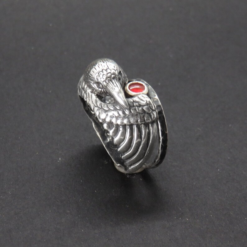 May include: A silver ring with a detailed bird design and a red gemstone. The bird's wings are spread out and the gemstone is set in the center of the bird's chest.