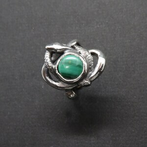 May include: A silver ring with a green malachite stone set in the center, surrounded by a design of two intertwined snakes.