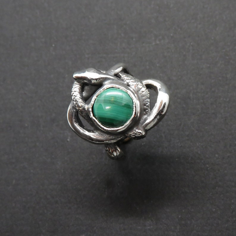 May include: A silver ring with a green malachite stone set in the center, surrounded by a design of two intertwined snakes.