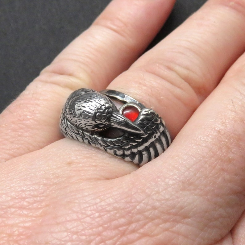 May include: A silver ring with a detailed bird design and a red gemstone in the eye. The bird's wings are spread out and the ring is worn on a finger.