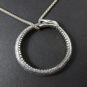 Può includere: Collana con ciondolo a forma di serpente in argento con un designo a serpente avvolto. La testa del serpente è sollevata e la coda è avvolta attorno al corpo del serpente per formare un cerchio.