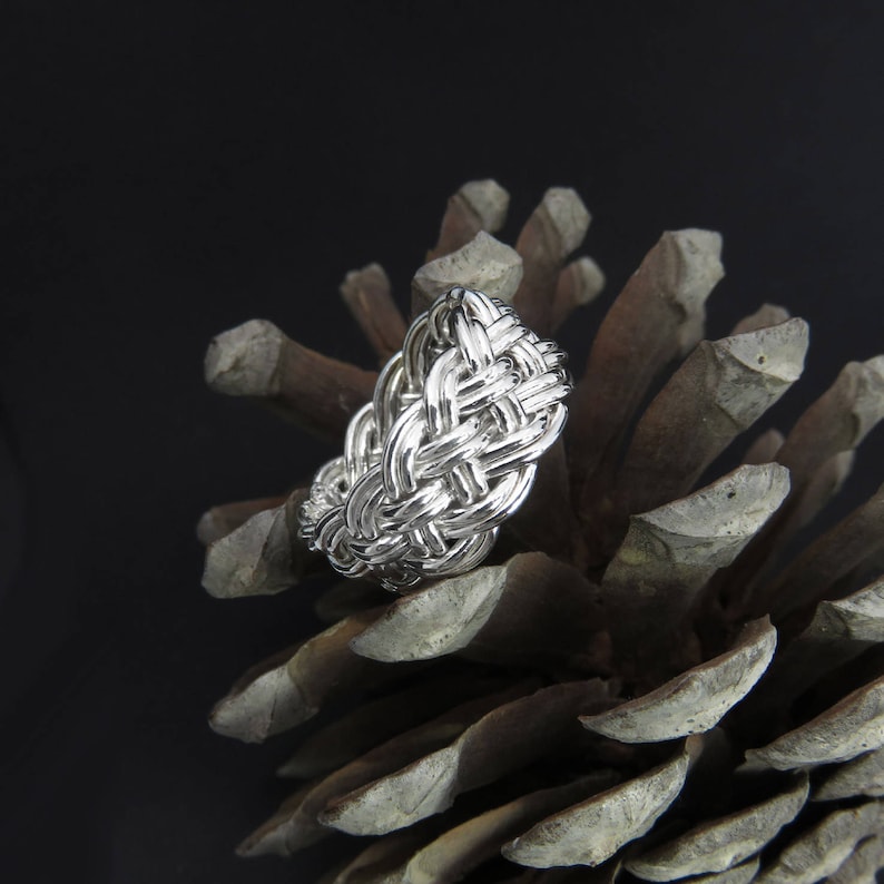 May include: A silver ring with a woven design, resting on a pine cone.