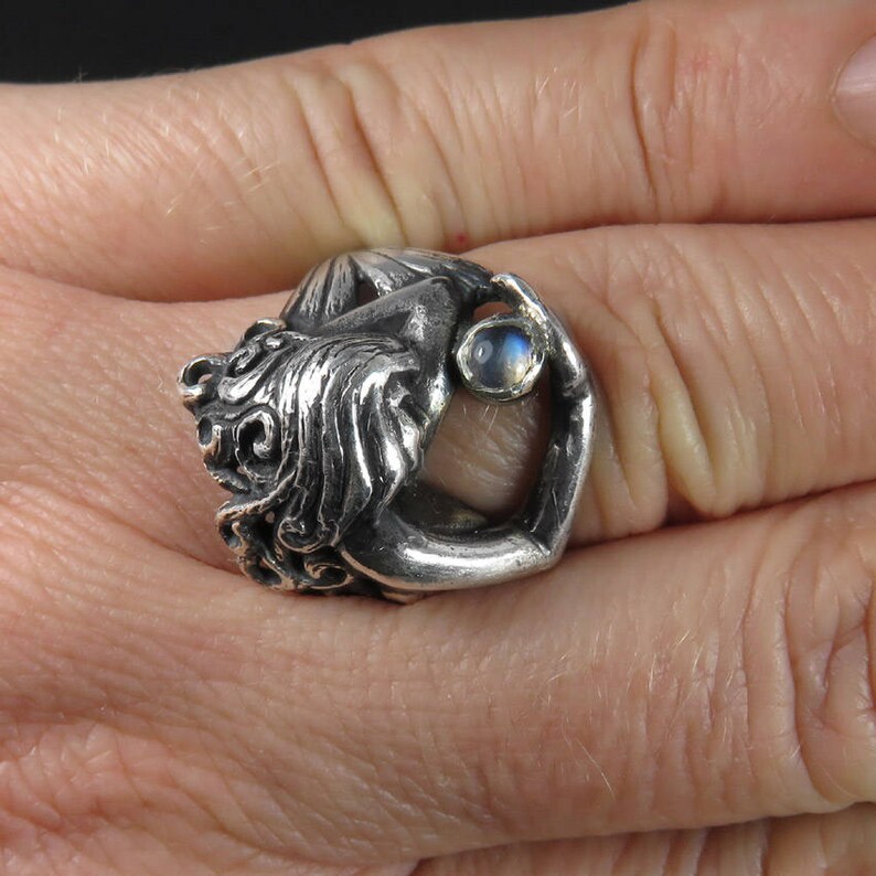 May include: A silver ring featuring a detailed sculpture of a woman holding a round iridescent gemstone.