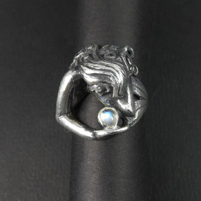 May include: A silver ring with a detailed sculpture of a woman holding a round iridescent gemstone.