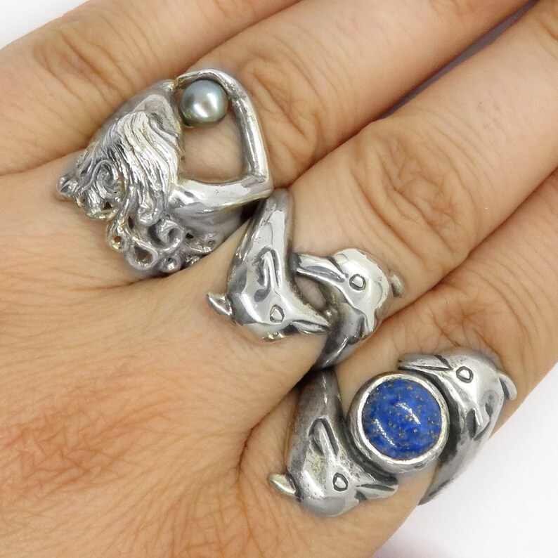 May include: Three silver rings featuring a mermaid holding a pearl, two dolphins intertwined, and two dolphins with a blue lapis lazuli stone.