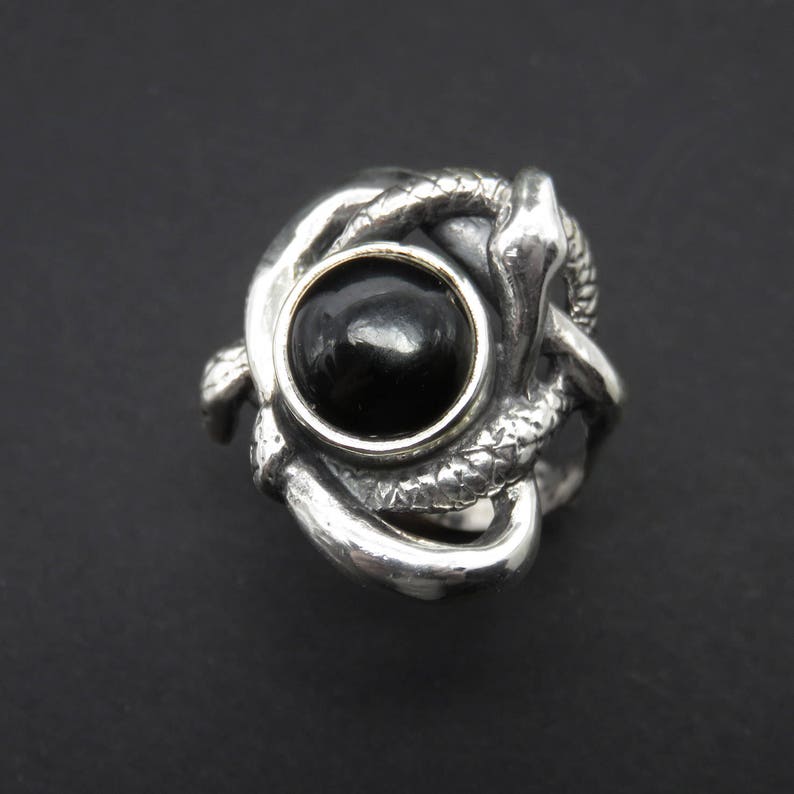 May include: A silver ring with a black gemstone set in a gold bezel. The ring features a design of two intertwined snakes.