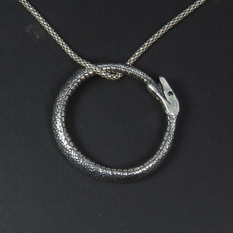 Op de afbeelding: Een zilveren ketting met een slangenhanger. De slang is opgerold tot een cirkel en zijn kop is naar voren gericht. De ketting is gemaakt van een delicate ketting.