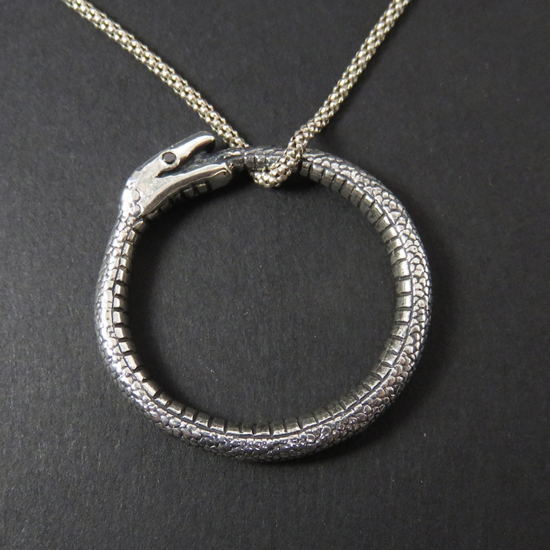 Op de afbeelding: Zilveren slangenhangerketting met een slang die een cirkel vormt. De slang is gedetailleerd met schubben en heeft een kleine zwarte steen in zijn oog.