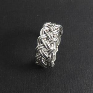 May include: A silver ring with a braided design. The ring is made of a single piece of metal that is woven together to create a complex pattern.