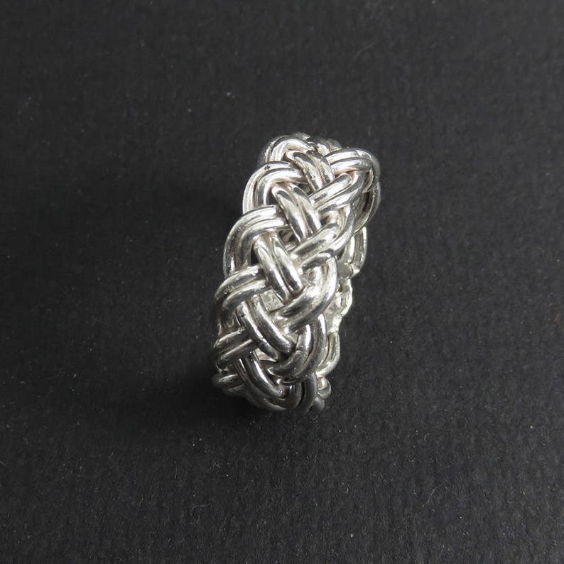 May include: A silver ring with a braided design. The ring is made of a single piece of metal that is woven together to create a complex pattern.