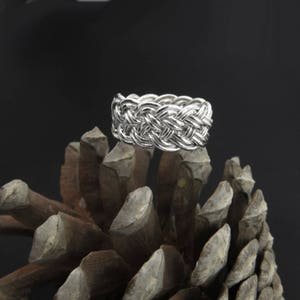 May include: A silver braided ring, with a woven design, is displayed on a brown pine cone.