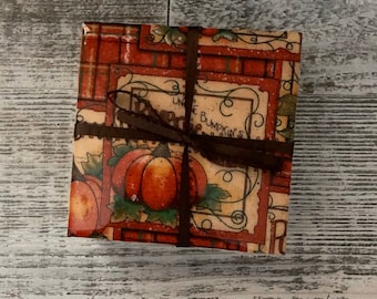 May include: A square gift box wrapped in paper with a fall theme. The paper features illustrations of pumpkins, leaves, and the text "Uncle Bumpkin's". A dark brown ribbon is tied around the box.