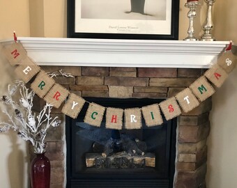May include: A burlap banner with the words "MERRY CHRISTMAS" in white, red, and green letters. The banner is strung across a fireplace mantel, with a stone fireplace in the background. Decorative items are on the mantel.