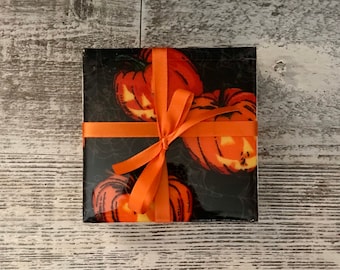 May include: A square set of coasters with a Halloween theme. The coasters feature orange jack-o'-lanterns on a black background with a spiderweb design. The set is tied with an orange ribbon, creating a festive presentation.