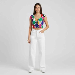 May include: A woman wearing a colorful patchwork crop top and white wide-leg jeans. The top features a V-neck and a vibrant mix of colors and patterns. The jeans are high-waisted and flow down to the ground.