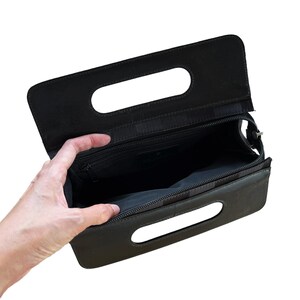 May include: Black clutch purse with a rectangular shape and two handle cutouts. The purse has a zipper closure and a black interior. The open purse reveals a small interior pocket. The purse is held open by a hand.
