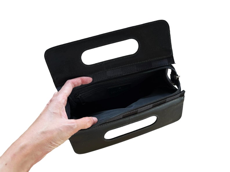 May include: Black clutch purse with a rectangular shape and two handle cutouts. The purse has a zipper closure and a black interior. The open purse reveals a small interior pocket. The purse is held open by a hand.