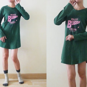 May include: A green long-sleeved dress with a pink graphic that says "ENJOY! BUBBLE" on the front. The dress has a relaxed fit and a slightly flared hem.
