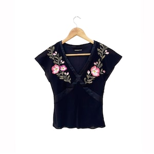 May include: Black cap-sleeve top with a V-neck and floral embroidery. The embroidery features pink flowers and green leaves. The top has a satin trim and is hanging on a wooden hanger.