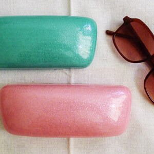 May include: Two rectangular glitter eyeglass cases, one teal green and one pink, are shown on a white fabric surface. A pair of brown sunglasses are also shown on the surface.
