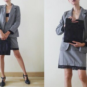 May include: A gray pinstripe blazer and skirt suit with black lace trim. The suit is paired with black heels and a black handbag. The outfit is worn by a person.