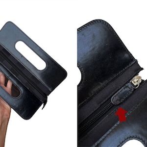 May include: Black leather clutch with two rectangular cut-out handles. The clutch has a zipper closure and a silver-toned zipper pull. The leather appears to be slightly worn. The clutch is held open, displaying the interior.