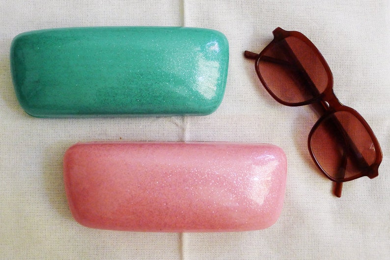 May include: Two rectangular glitter eyeglass cases, one teal green and one pink, are shown on a white fabric surface. A pair of brown sunglasses are also shown on the surface.