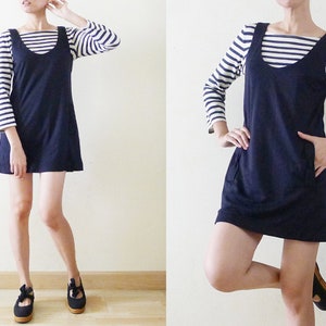 May include: A navy blue sleeveless dress with a scoop neck and two pockets. The dress is worn over a white and navy striped long-sleeved shirt.