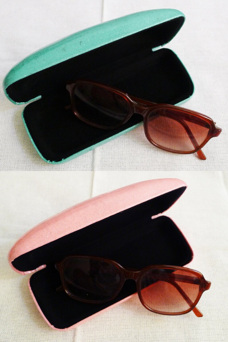 May include: Two pairs of sunglasses in their cases. The cases are open and the sunglasses are inside. The case on the top is teal and the case on the bottom is pink.