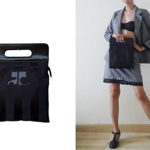 May include: Black tote bag with a handle and a rectangular shape. The bag has vertical stripes and a logo on the front. The top handle is black and shiny. The bag is shown next to a person wearing a gray suit.