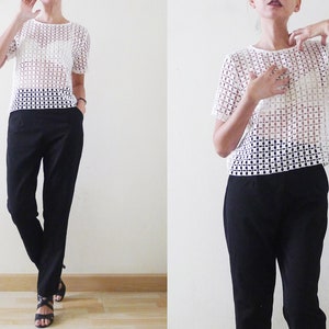 May include: A white crochet top with a square pattern and short sleeves. The top is worn over a white bra and paired with black pants.