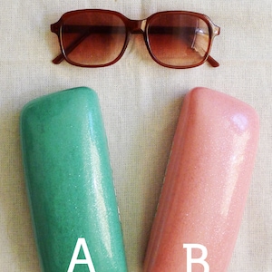 May include: Two rectangular eyeglass cases, one green and one pink, with the letters A and B printed on them. The cases are made of a sparkly material. A pair of brown sunglasses are in the background.