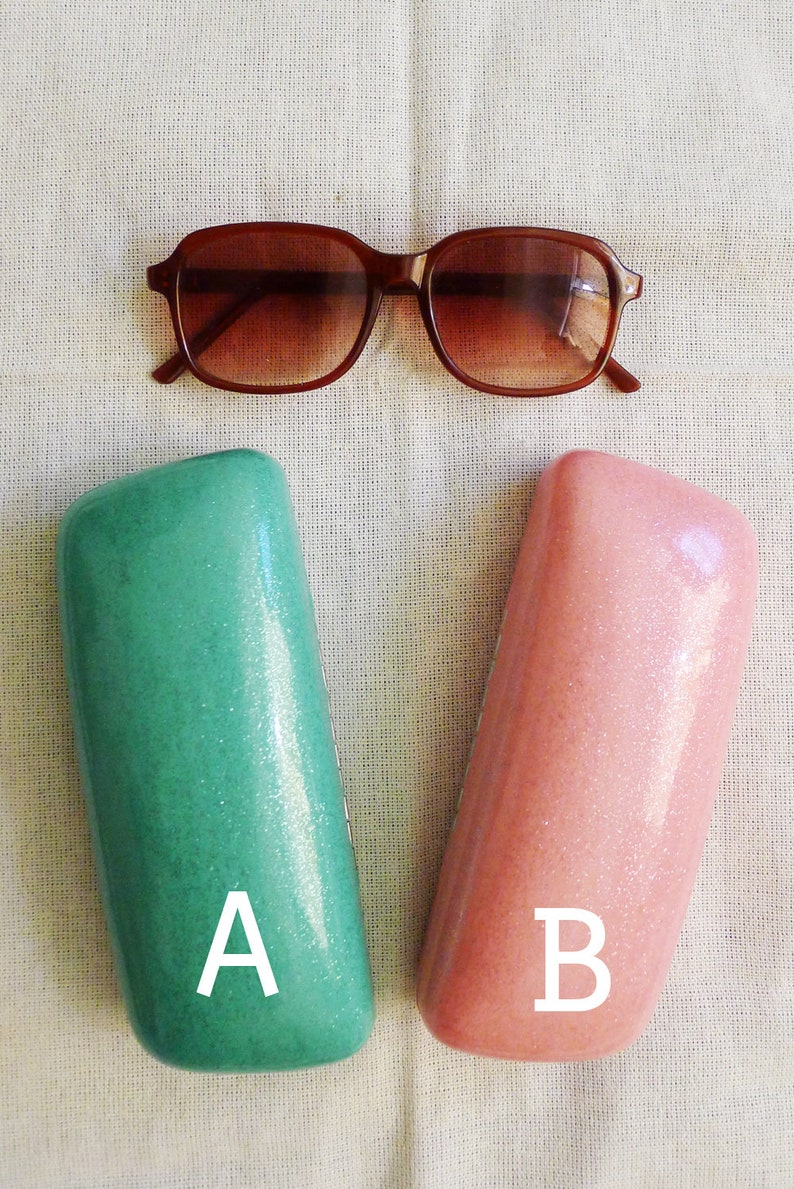 May include: Two rectangular eyeglass cases, one green and one pink, with the letters A and B printed on them. The cases are made of a sparkly material. A pair of brown sunglasses are in the background.