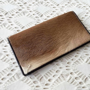 Puede incluir: Una cartera rectangular con una textura similar a la piel de color marrón y beige. La cartera tiene un borde negro y está sobre un fondo blanco estampado. La cartera parece estar cerrada.