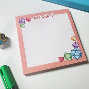 May include: A pink notepad with a patterned border and the text "Roll with it!" features colorful dice illustrations. A blue and gold die and a green pen are also visible.