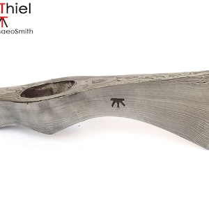 Op de afbeelding: Een handgesmeed bijlkop met een damaststaalpatroon. De bijlkop heeft een gebogen vorm en een gat voor een steel. De bijlkop is gemarkeerd met de letter "T" in een cirkel.