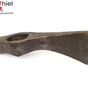 Puede incluir: Una cabeza de hacha de acero forjada a mano con una hoja curva y un agujero para un mango. La cabeza del hacha es marrón y tiene una marca "AT" en el lateral.