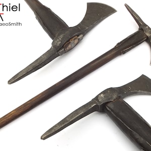 May include: Three views of a medieval style axe with a wooden handle. The axe has a pick on one side and a blade on the other. The axe is made of metal and has a dark brown wooden handle. The axe is displayed on a white background. AdiThiel the Archaeosmith is printed on the background.
