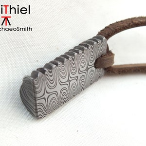 Könnte beinhalten: Ein silberner Anhänger mit einem gemusterten Design und einer braunen Lederschnur. Der Anhänger hat einen gezackten Rand und ist rechteckig.