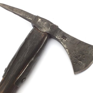 May include: A close-up of a medieval axe with a wooden handle and a metal head. The axe has a sharp blade and a pointed spike on the back of the head. The handle is wrapped in leather and has a metal band around it. The axe is a dark gray color and has a worn appearance.
