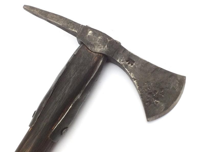 May include: A close-up of a medieval axe with a wooden handle and a metal head. The axe has a sharp blade and a pointed spike on the back of the head. The handle is wrapped in leather and has a metal band around it. The axe is a dark gray color and has a worn appearance.