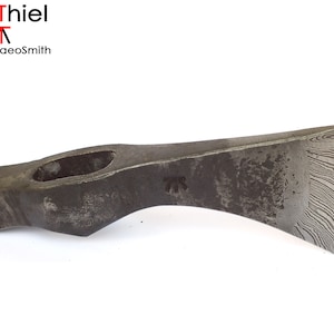 Op de afbeelding: Een handgesmeed bijlkop met een lemmet van damaststaal. De bijlkop heeft een gat voor een steel en een opvallend patroon op het lemmet.