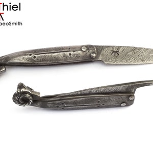 Może przedstawiać: Ręcznie kuty, składany nóż z damaszkowej stali z unikalnym, ozdobnym uchwytem. Ostrze jest otwarte, a uchwyt zamknięty.